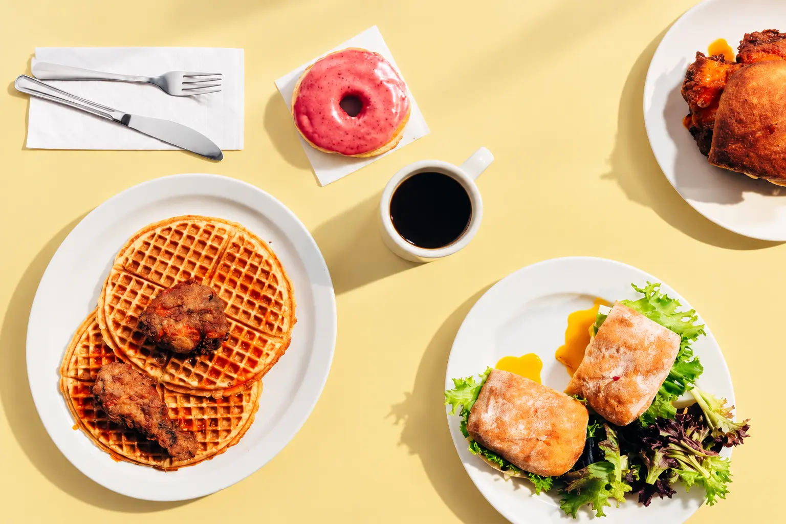 Gracie’s waffles, coffee, a pink glazed donut, and a breakfast sandwich on a butter-yellow table